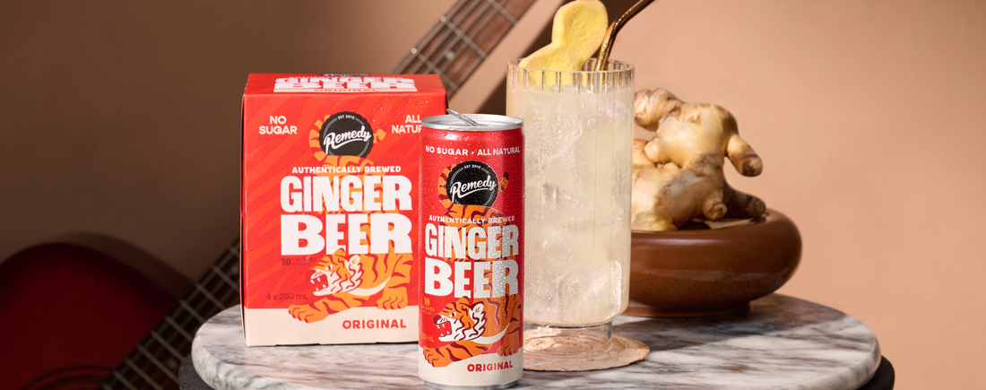 A 4-pack of Remedy Ginger Beer with a can standing in front of it. Next to the can there's a glass filled with Ginger Beer, alongside a piece of ginger.